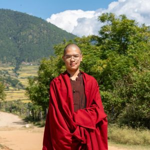 Monk in traditional red robes standing in a serene mountain landscape under a clear blue sky.